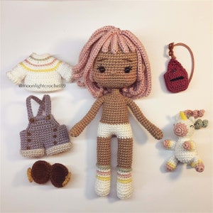 Puede incluir: Una mu&ntilde;eca de crochet con pelo rosa, piel morena y un vestido blanco. La mu&ntilde;eca sostiene un peque&ntilde;o unicornio de crochet. La mu&ntilde;eca lleva un mono azul y un jersey a rayas. La mu&ntilde;eca tambi&eacute;n lleva un par de calcetines blancos con rayas rosas y blancas.