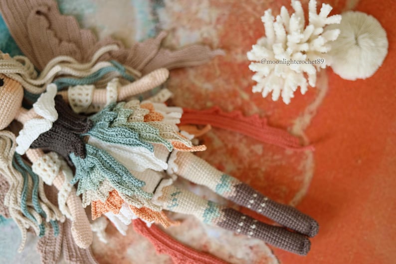Puede incluir: Una mu&ntilde;eca hada de ganchillo hecha a mano con cabello largo y ondulado, un corpi&ntilde;o marr&oacute;n oscuro y una falda en capas con detalles en forma de hoja en blanco, verde azulado y naranja. La mu&ntilde;eca incluye el texto @moonlightcrochet89.
