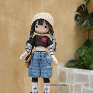 May include: A crocheted doll wearing a beige beanie, a brown and white striped shirt, blue denim shorts with pockets, and red and white sneakers. The doll has brown hair, blue eyes, and a red heart on its shirt.