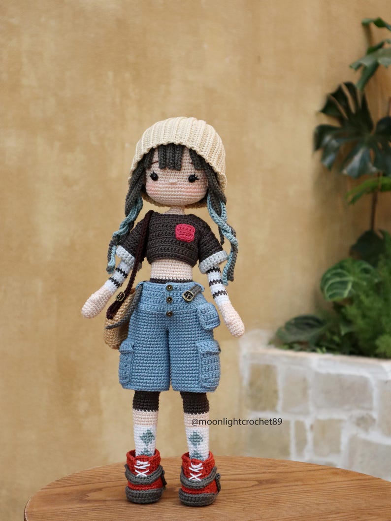 May include: A crocheted doll wearing a beige beanie, a brown and white striped shirt, blue denim shorts with pockets, and red and white sneakers. The doll has brown hair, blue eyes, and a red heart on its shirt.