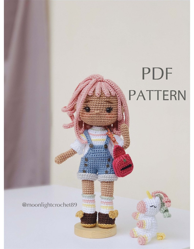 Puede incluir: Patr&oacute;n de mu&ntilde;eca de crochet en PDF. La mu&ntilde;eca lleva un vestido de mezclilla azul con una camisa blanca y cabello rosado. Lleva una bolsa roja y est&aacute; de pie junto a un unicornio blanco.
