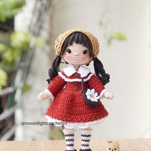 May include: A crocheted doll with brown hair, wearing a red dress, white socks, and black shoes. The doll is holding a small crocheted purse with a white flower. The doll is standing next to a small crocheted teddy bear.