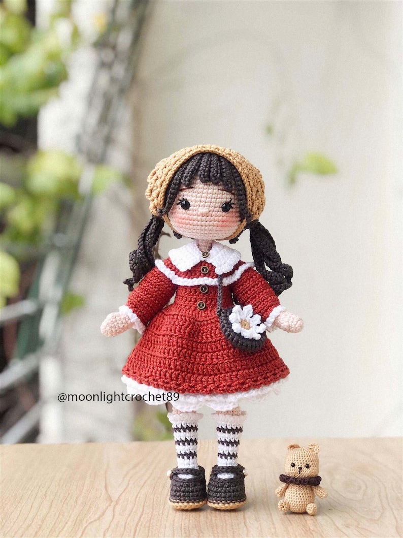 May include: A crocheted doll with brown hair, wearing a red dress, white socks, and black shoes. The doll is holding a small crocheted purse with a white flower. The doll is standing next to a small crocheted teddy bear.