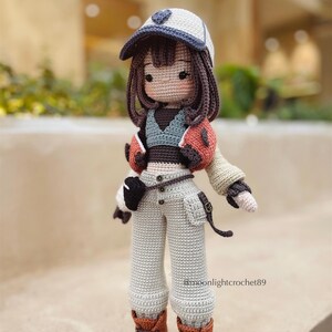 Puede incluir: Una mu&ntilde;eca de crochet que lleva una gorra de b&eacute;isbol blanca y azul, una chaqueta marr&oacute;n y naranja, un top verde y marr&oacute;n, pantalones beige y botas marrones. La mu&ntilde;eca tiene el pelo marr&oacute;n y un dise&ntilde;o en forma de coraz&oacute;n en la gorra.