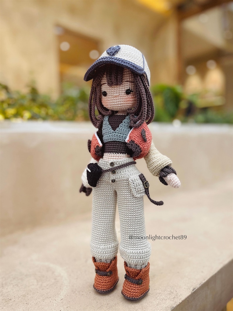 Puede incluir: Una mu&ntilde;eca de crochet que lleva una gorra de b&eacute;isbol blanca y azul, una chaqueta marr&oacute;n y naranja, un top verde y marr&oacute;n, pantalones beige y botas marrones. La mu&ntilde;eca tiene el pelo marr&oacute;n y un dise&ntilde;o en forma de coraz&oacute;n en la gorra.