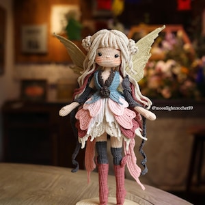 Peut inclure: Poup&eacute;e de f&eacute;e au crochet faite &agrave; la main, avec de longs cheveux blonds, de grandes ailes et une robe &agrave; plusieurs &eacute;paisseurs dans les tons de rose, de bleu et de cr&egrave;me. La poup&eacute;e est pos&eacute;e sur une base en bois. Les bottes de la poup&eacute;e sont de couleur rose poudr&eacute;.