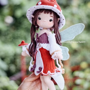 May include: A handmade crochet fairy doll with a red and white mushroom cap hat, long brown hair, and light blue wings. The doll wears a red and white dress with a purple bodice. The text "@moonlightcrochet89" is visible.