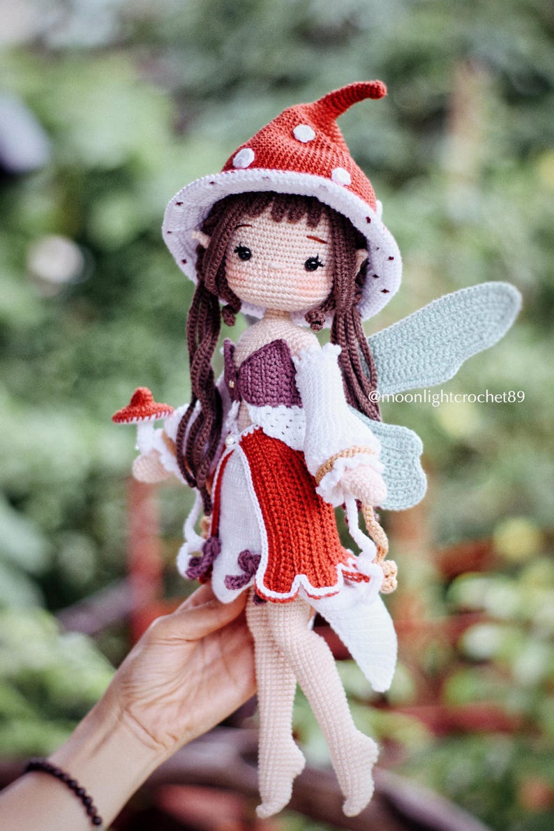 May include: A handmade crochet fairy doll with a red and white mushroom cap hat, long brown hair, and light blue wings. The doll wears a red and white dress with a purple bodice. The text "@moonlightcrochet89" is visible.