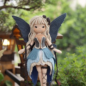 May include: A handmade crochet fairy doll with long blonde hair, blue wings, and a blue and tan dress. The doll is standing on a wooden slice, with a natural green background. The doll has a delicate design and is a unique handmade item.