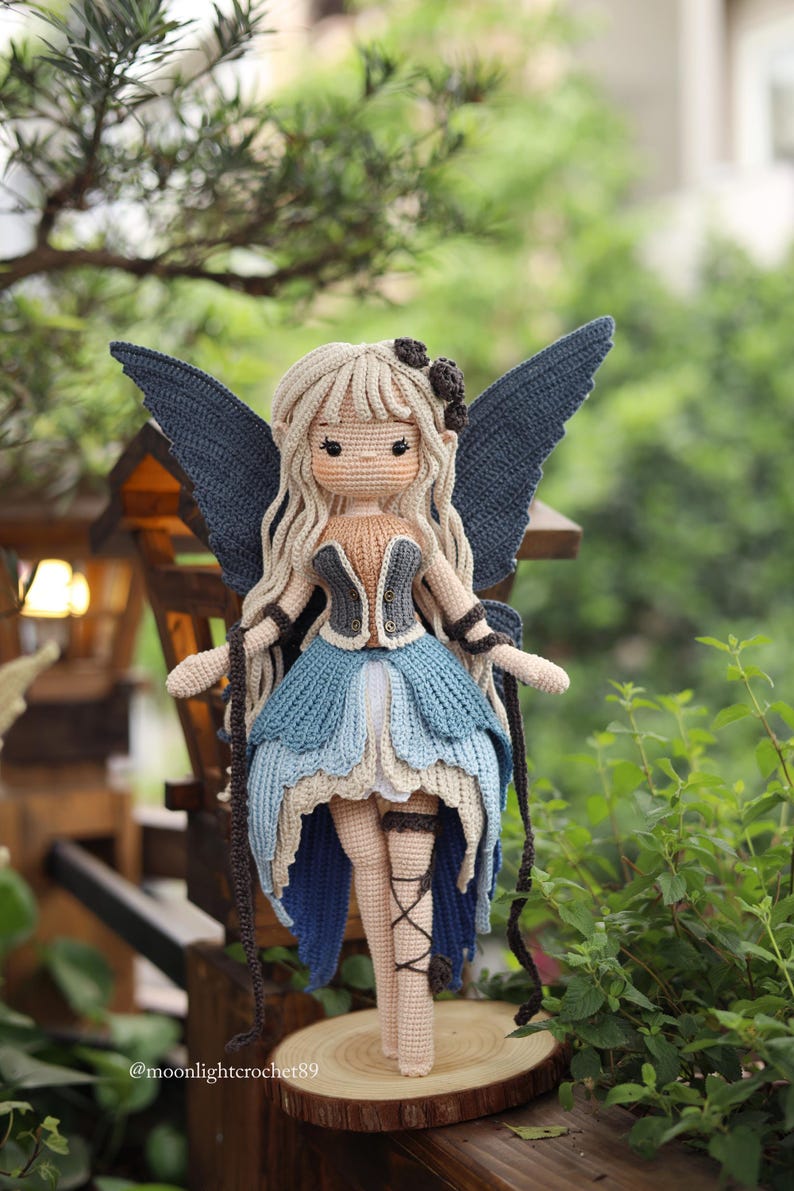 May include: A handmade crochet fairy doll with long blonde hair, blue wings, and a blue and tan dress. The doll is standing on a wooden slice, with a natural green background. The doll has a delicate design and is a unique handmade item.