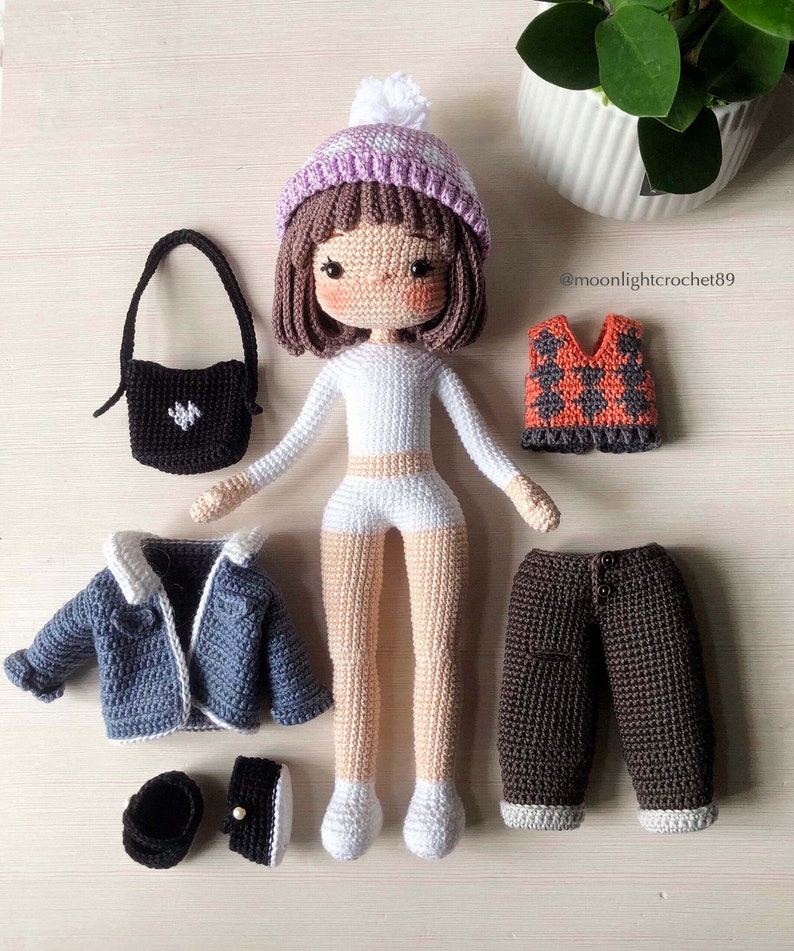 May include: Crocheted doll with brown hair wearing a purple and white hat, white shirt, white shorts, and brown trousers. The doll is holding a black handbag. The doll is surrounded by other crocheted clothing items, including a blue jacket, a brown waistcoat, and a pair of brown shoes. The image includes the text "@moonlightcrochet89".