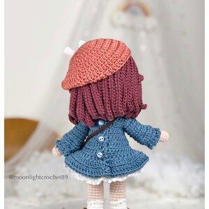 May include: A crocheted doll wearing a blue dress, a pink beret, and brown shoes. The doll has long, dark pink hair and is standing on a wooden base.