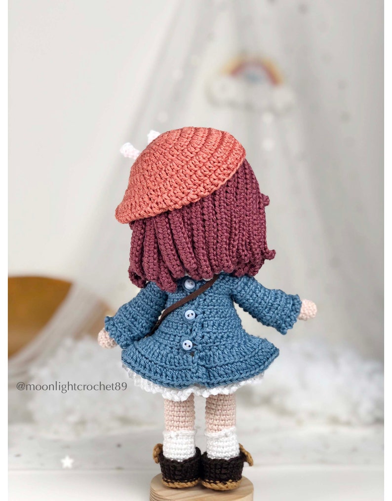 May include: A crocheted doll wearing a blue dress, a pink beret, and brown shoes. The doll has long, dark pink hair and is standing on a wooden base.