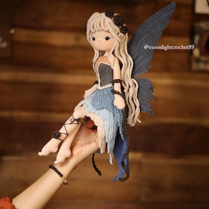 May include: A handmade crochet fairy doll with long blonde hair, blue wings, and a blue and beige dress. The doll has a detailed design with a dark bodice and is held in a hand. The doll's legs are adorned with black accents. The doll is a unique, handcrafted item.