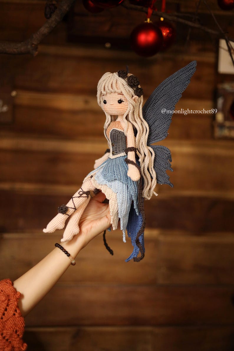 May include: A handmade crochet fairy doll with long blonde hair, blue wings, and a blue and beige dress. The doll has a detailed design with a dark bodice and is held in a hand. The doll's legs are adorned with black accents. The doll is a unique, handcrafted item.