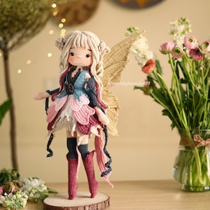 Peut inclure: Une poup&eacute;e de f&eacute;e au crochet faite &agrave; la main avec des cheveux blonds, portant une robe color&eacute;e avec des accents roses, bleus et blancs. La poup&eacute;e a des ailes dor&eacute;es et se tient sur une base en bois. Un vase de fleurs est en arri&egrave;re-plan.