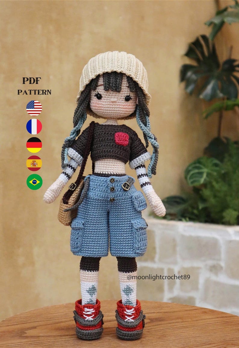 May include: Crocheted doll with blue hair, wearing a beige beanie, black and white striped shirt, blue denim shorts, and red and gray shoes. The doll is holding a brown bag. The text "PDF PATTERN" and flags of the United States, France, Germany, Spain, and Brazil are visible in the background. The text "@moonlightcrochet89" is visible in the bottom right corner.
