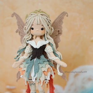 Puede incluir: Una mu&ntilde;eca de ganchillo hecha a mano con pelo largo rubio, ojos grandes y oscuros y tez bronceada. La mu&ntilde;eca lleva un corpi&ntilde;o marr&oacute;n oscuro, una falda a capas en tonos verde, blanco y naranja, y grandes alas marrones. El texto dice @moonlightcrochet89.