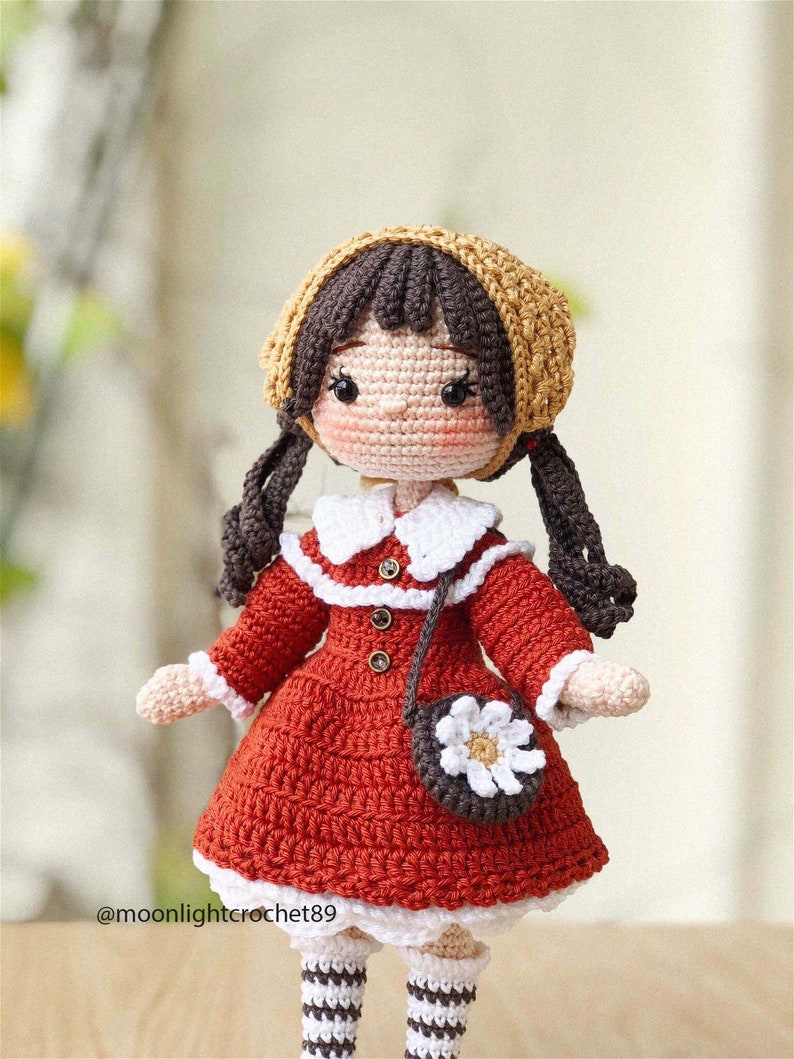 May include: A crocheted doll with brown hair, wearing a red dress, a white collar, a brown purse with a white flower, and a brown and yellow hat. The doll is wearing white and brown striped socks.