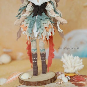 Puede incluir: Una mu&ntilde;eca de ganchillo hecha a mano con cabello largo y fluido, un corpi&ntilde;o marr&oacute;n oscuro y una falda en capas en tonos verde salvia, crema y naranja. La mu&ntilde;eca lleva botas altas marrones y tiene el texto @moonlightcrochet89 impreso al lado.