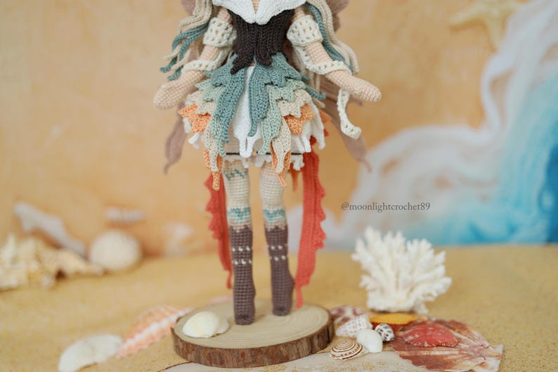 Puede incluir: Una mu&ntilde;eca de ganchillo hecha a mano con cabello largo y fluido, un corpi&ntilde;o marr&oacute;n oscuro y una falda en capas en tonos verde salvia, crema y naranja. La mu&ntilde;eca lleva botas altas marrones y tiene el texto @moonlightcrochet89 impreso al lado.