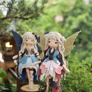 May include: Two handmade crochet fairy dolls with detailed wings and layered skirts. One doll wears a blue and black outfit, the other a pink, white, and red outfit. Both dolls have blonde hair and are displayed on wooden bases.