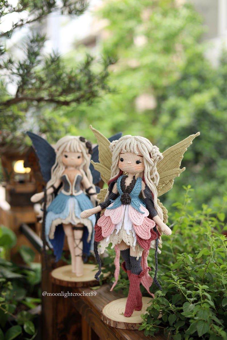 May include: Two handmade crochet fairy dolls with detailed wings and layered skirts. One doll wears a blue and black outfit, the other a pink, white, and red outfit. Both dolls have blonde hair and are displayed on wooden bases.