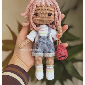 Puede incluir: Una mu&ntilde;eca de crochet con pelo rosa, que lleva una camisa a rayas azules y blancas, un mono azul y calcetines blancos con rayas rosas, amarillas y blancas. La mu&ntilde;eca lleva un peque&ntilde;o bolso rojo de crochet.