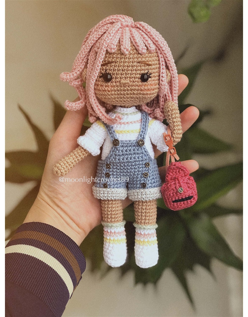Puede incluir: Una mu&ntilde;eca de crochet con pelo rosa, que lleva una camisa a rayas azules y blancas, un mono azul y calcetines blancos con rayas rosas, amarillas y blancas. La mu&ntilde;eca lleva un peque&ntilde;o bolso rojo de crochet.