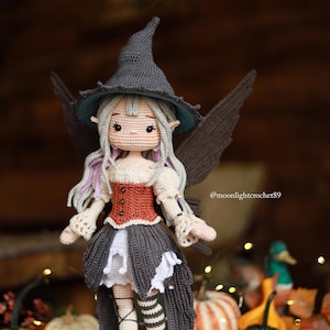 May include: A handmade crochet doll dressed as a fairy witch. The doll wears a gray witch hat, a red and cream bodice, a gray skirt, and striped stockings. The doll has gray hair with purple highlights and black wings. The background is blurred with pumpkins.