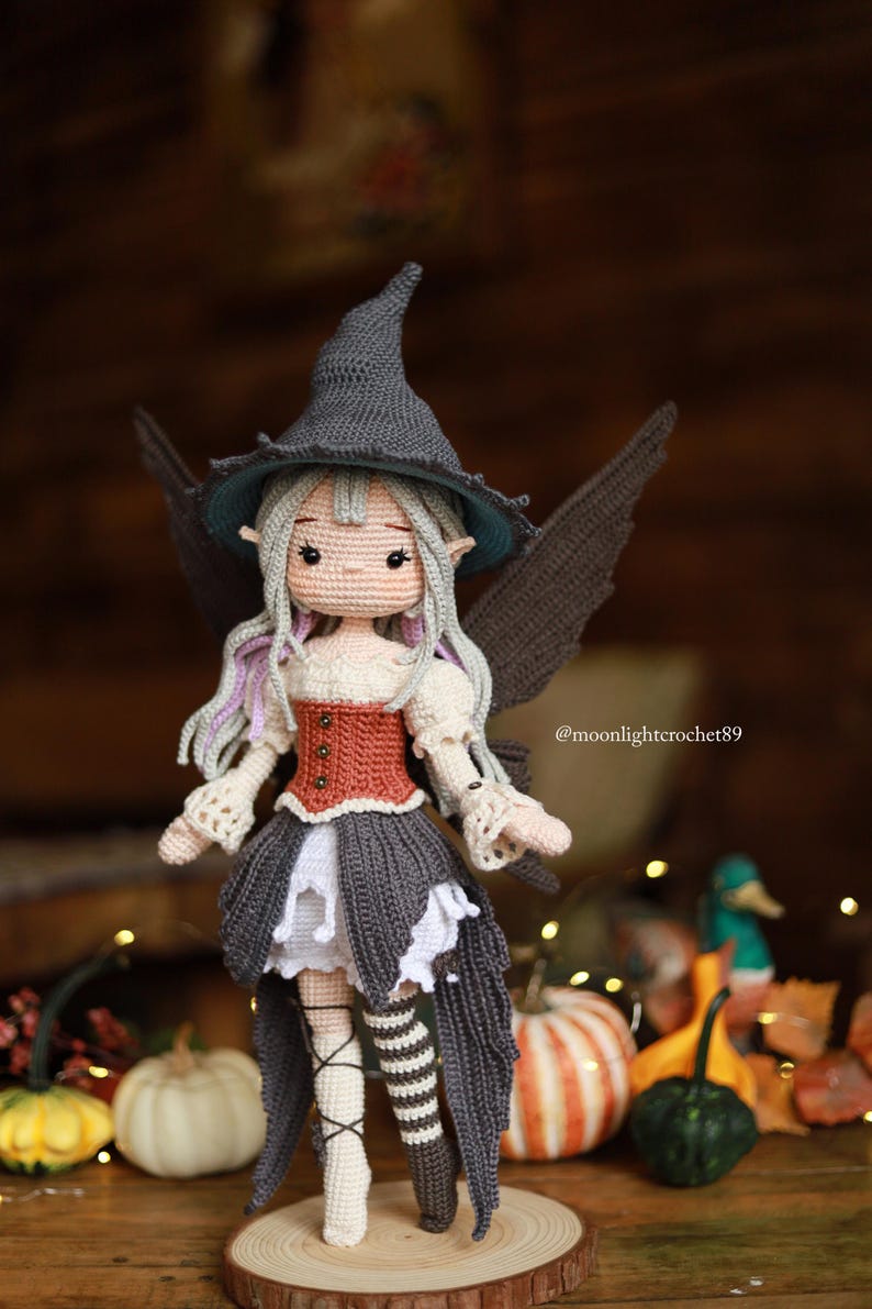 May include: A handmade crochet doll dressed as a fairy witch. The doll wears a gray witch hat, a red and cream bodice, a gray skirt, and striped stockings. The doll has gray hair with purple highlights and black wings. The background is blurred with pumpkins.
