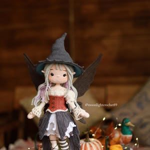 May include: A handmade crochet doll of a fairy witch, wearing a gray pointed hat, a red and white bodice, and a gray skirt. The doll has gray wings and is standing on a wooden base. The doll is surrounded by pumpkins and fall foliage.