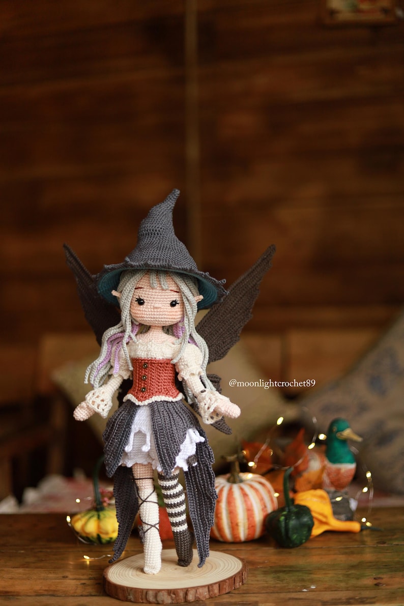 May include: A handmade crochet doll of a fairy witch, wearing a gray pointed hat, a red and white bodice, and a gray skirt. The doll has gray wings and is standing on a wooden base. The doll is surrounded by pumpkins and fall foliage.