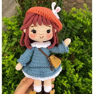 May include: A crocheted doll with brown hair, wearing a blue dress, a brown bag, a white crocheted hat with a white bunny ear, and brown shoes.