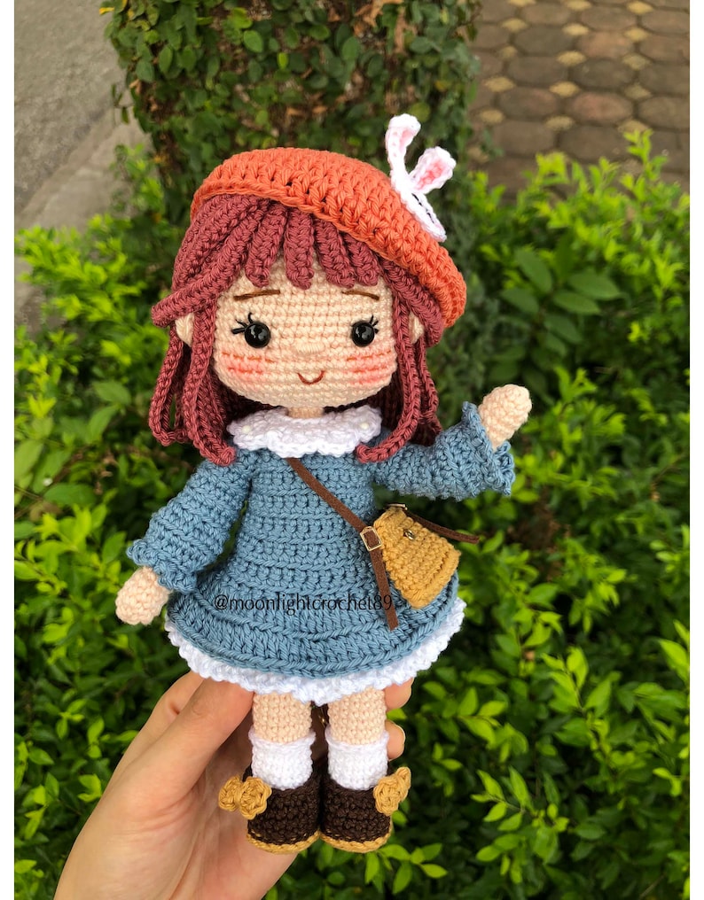 May include: A crocheted doll with brown hair, wearing a blue dress, a brown bag, a white crocheted hat with a white bunny ear, and brown shoes.