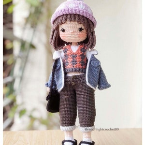 May include: Crocheted doll wearing a purple and white hat, a blue denim jacket, a red and white plaid waistcoat, brown trousers, and black shoes. The doll is holding a black bag.
