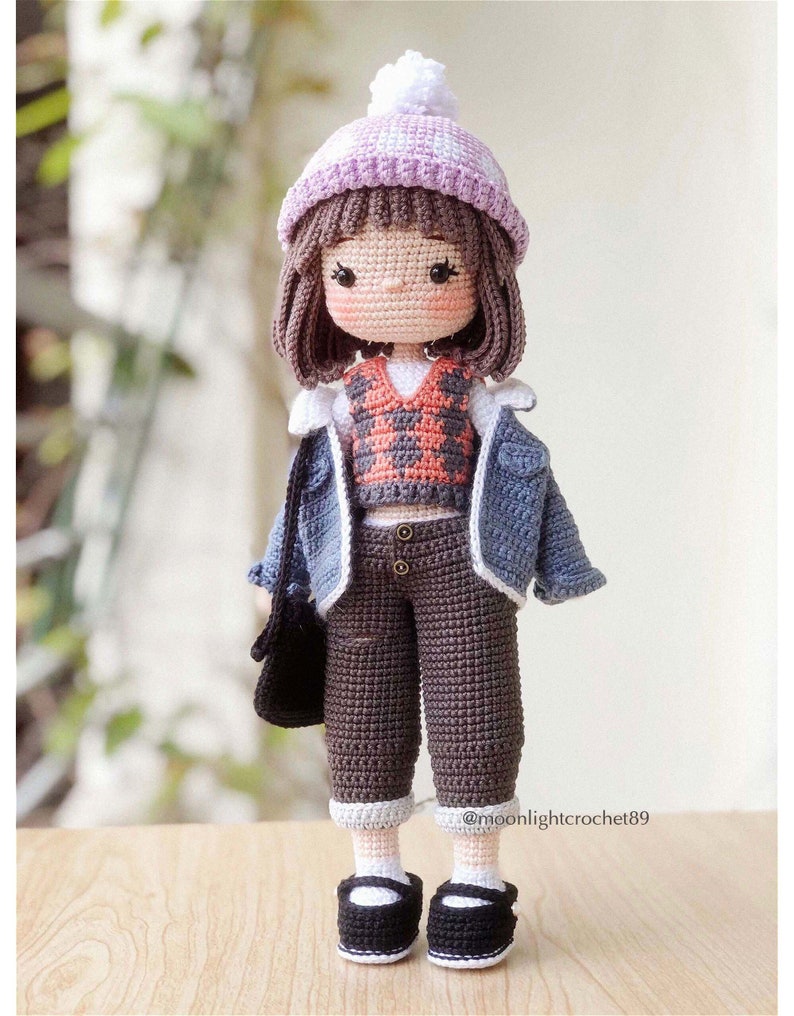 May include: Crocheted doll wearing a purple and white hat, a blue denim jacket, a red and white plaid waistcoat, brown trousers, and black shoes. The doll is holding a black bag.