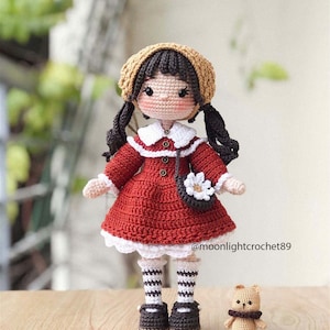 May include: A crocheted doll wearing a red dress, white socks, and black shoes. The doll has brown hair, a white collar, and a brown hat. The doll is holding a white flower purse. A small crocheted brown bear is sitting on the table next to the doll. @moonlightcrochet89