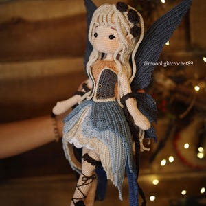 May include: A handmade crochet fairy doll with a light skin tone, blonde hair, and blue wings. The doll wears a blue and beige dress with black accents. The doll's legs are adorned with black lace-up details. The doll is a whimsical, handcrafted item.
