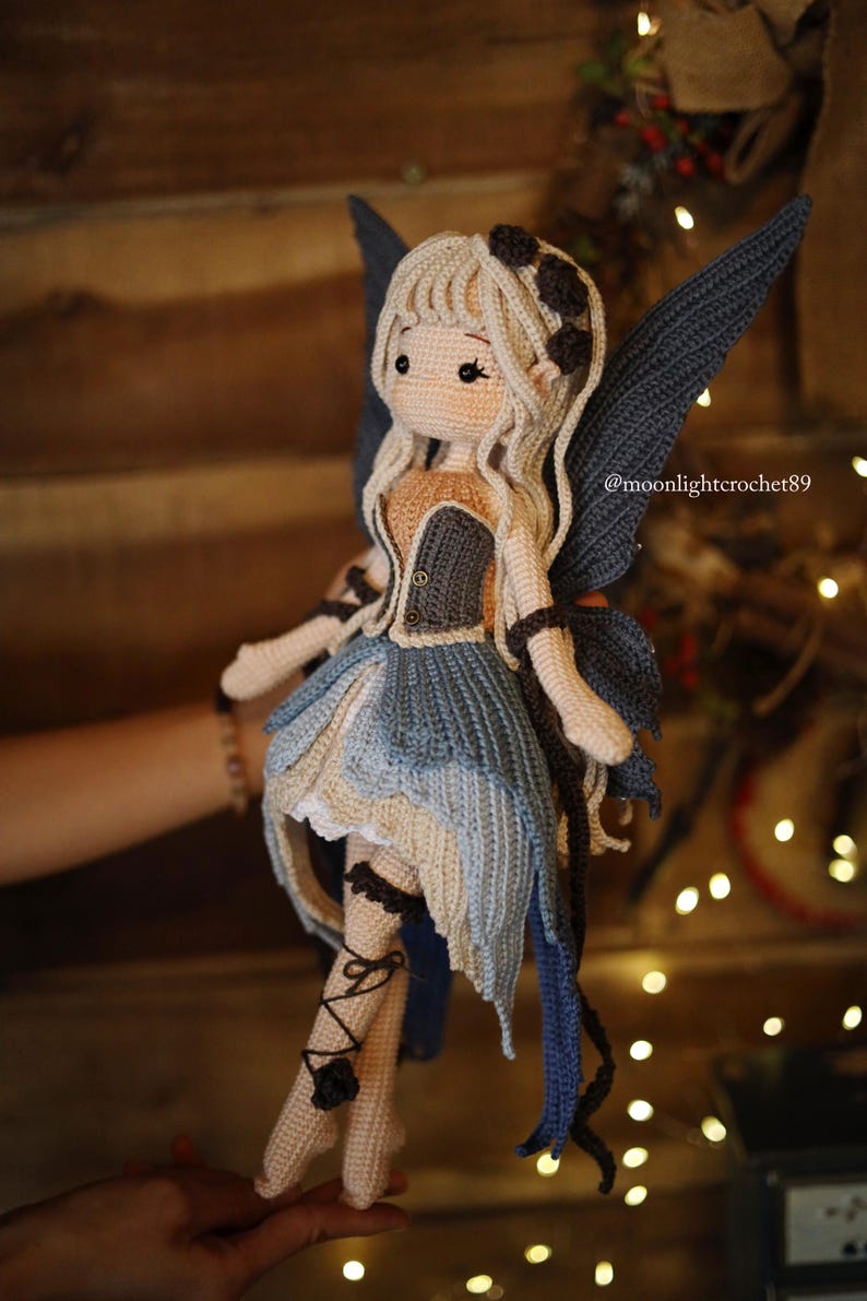 May include: A handmade crochet fairy doll with a light skin tone, blonde hair, and blue wings. The doll wears a blue and beige dress with black accents. The doll's legs are adorned with black lace-up details. The doll is a whimsical, handcrafted item.