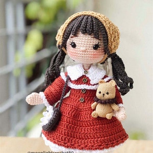 May include: A crocheted doll wearing a red dress, white collar, and brown hat. The doll has brown hair in pigtails and is holding a small crocheted teddy bear. The doll is wearing white and black striped socks.