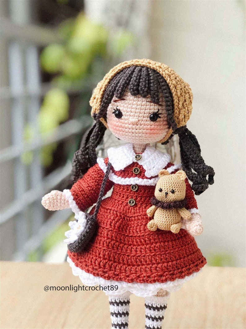 May include: A crocheted doll wearing a red dress, white collar, and brown hat. The doll has brown hair in pigtails and is holding a small crocheted teddy bear. The doll is wearing white and black striped socks.