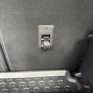 Op de afbeelding: Een close-up van een zwart auto-interieur met een "TOP TETHER" ankerpunt. Het anker is een rechthoekig, zilverkleurig metalen onderdeel met een vergrendeling. Daaronder een zwarte plastic opbergbak met een geometrisch patroon.