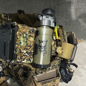 Könnte beinhalten: Eine beige und schwarze taktische Ausrüstungstasche mit einer schwarzen und grauen Wasserflasche mit einem Totenkopf- und Schlangenmotiv. Die Tasche hat einen schwarzen Gurt und eine schwarze Schnalle.