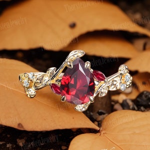 Op de afbeelding: Een goudkleurige ring met een grote, peervormige, dieprode edelsteen als middelpunt. De ring heeft een blad- en wijnstokontwerp met kleinere rode en heldere edelstenen. De ring is geplaatst tegen een achtergrond van herfstbladeren.