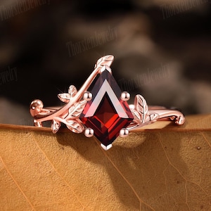 Könnte beinhalten: Ring aus Roségold mit einem rautenförmigen, tiefroten Edelstein. Der Ring hat ein zartes, von der Natur inspiriertes Design mit Blatt- und Ranken-Akzenten. Der Ring liegt auf einem braunen Blatt.