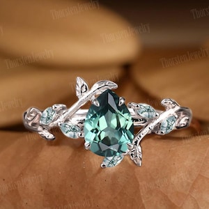 May include: A pear-shaped green gemstone ring with a silver band and delicate leaf accents. The ring is set on a brown background.