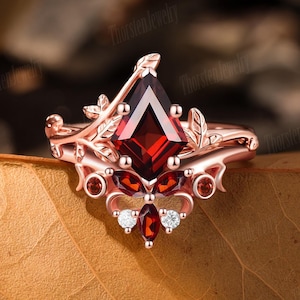 Könnte beinhalten: Roségoldener Ring mit einem großen, rautenförmigen, tiefroten Edelstein als Mittelpunkt. Der Ring hat kleinere rote und weiße Edelsteine sowie blattartige Details. Der Markenname "Thorsten Jewelry" ist sichtbar.