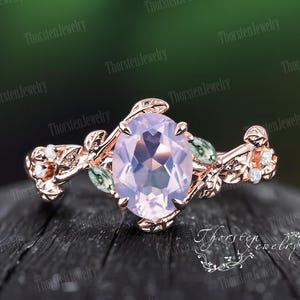 Peut inclure: Bague en or rose ornée d'une pierre ovale de couleur violet clair. La bague présente un motif floral avec des feuilles et de petits accents blancs. La bague est posée sur une surface en bois sombre avec un arrière-plan vert flou.