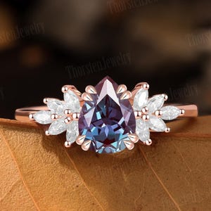 May include: A rose gold ring with a pear-shaped alexandrite gemstone that changes colour. The ring is surrounded by marquise-cut white gemstones. The ring is set against a brown leaf background. The text "Thorsten Jewelry" is visible.